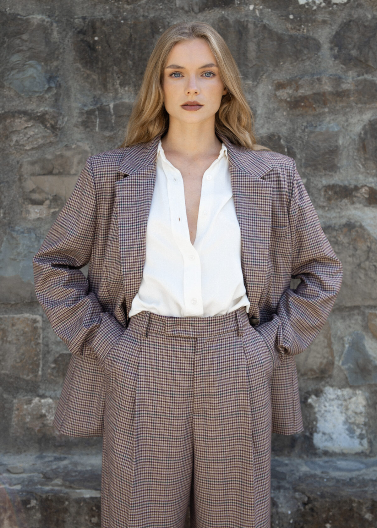 Model wearing brown checked wide-leg trousers with pleats and high waist, styled with matching blazer, photographed outdoors against a stone wall.