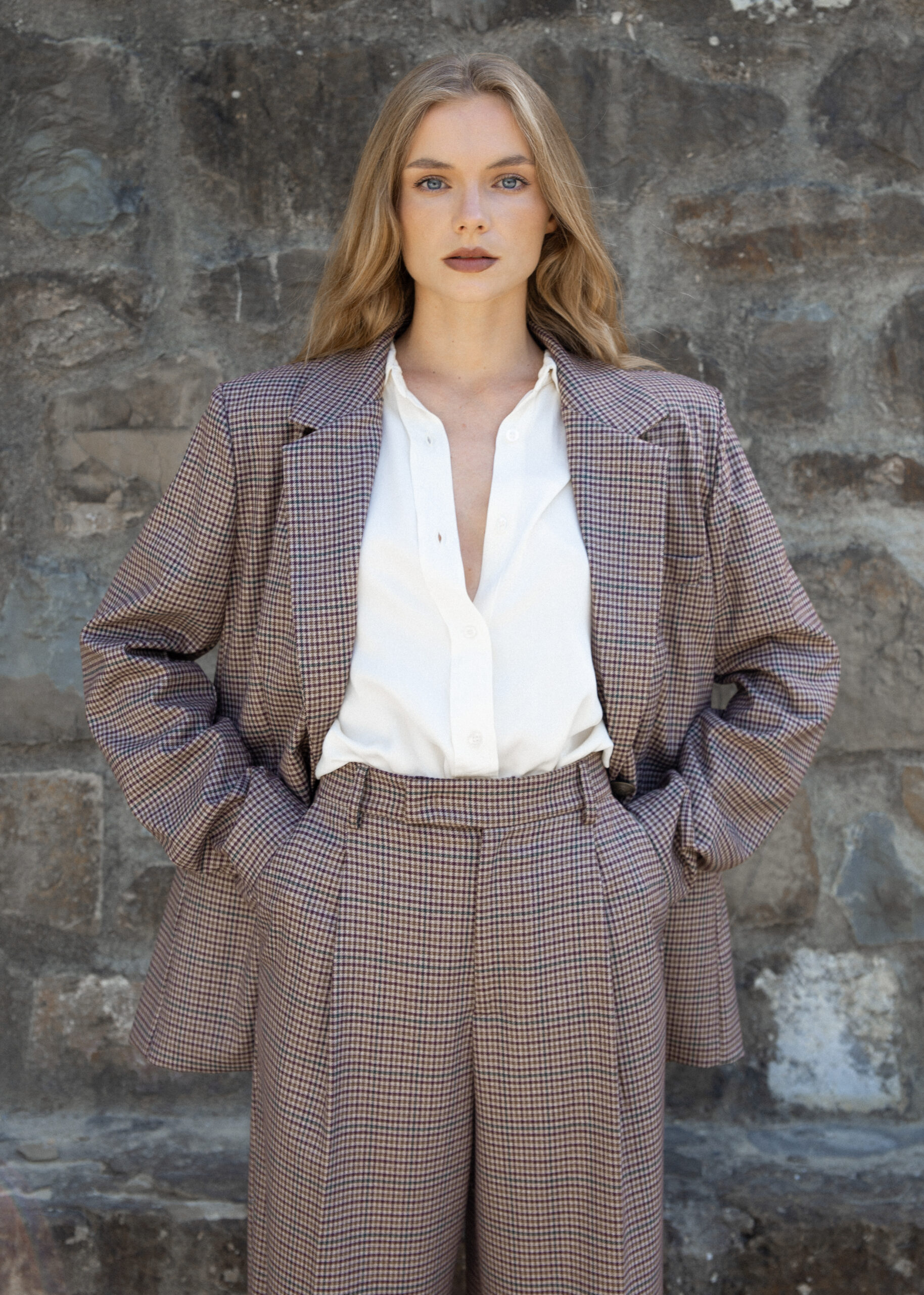 Model wearing brown checked wide-leg trousers with pleats and high waist, styled with matching blazer, photographed outdoors against a stone wall.