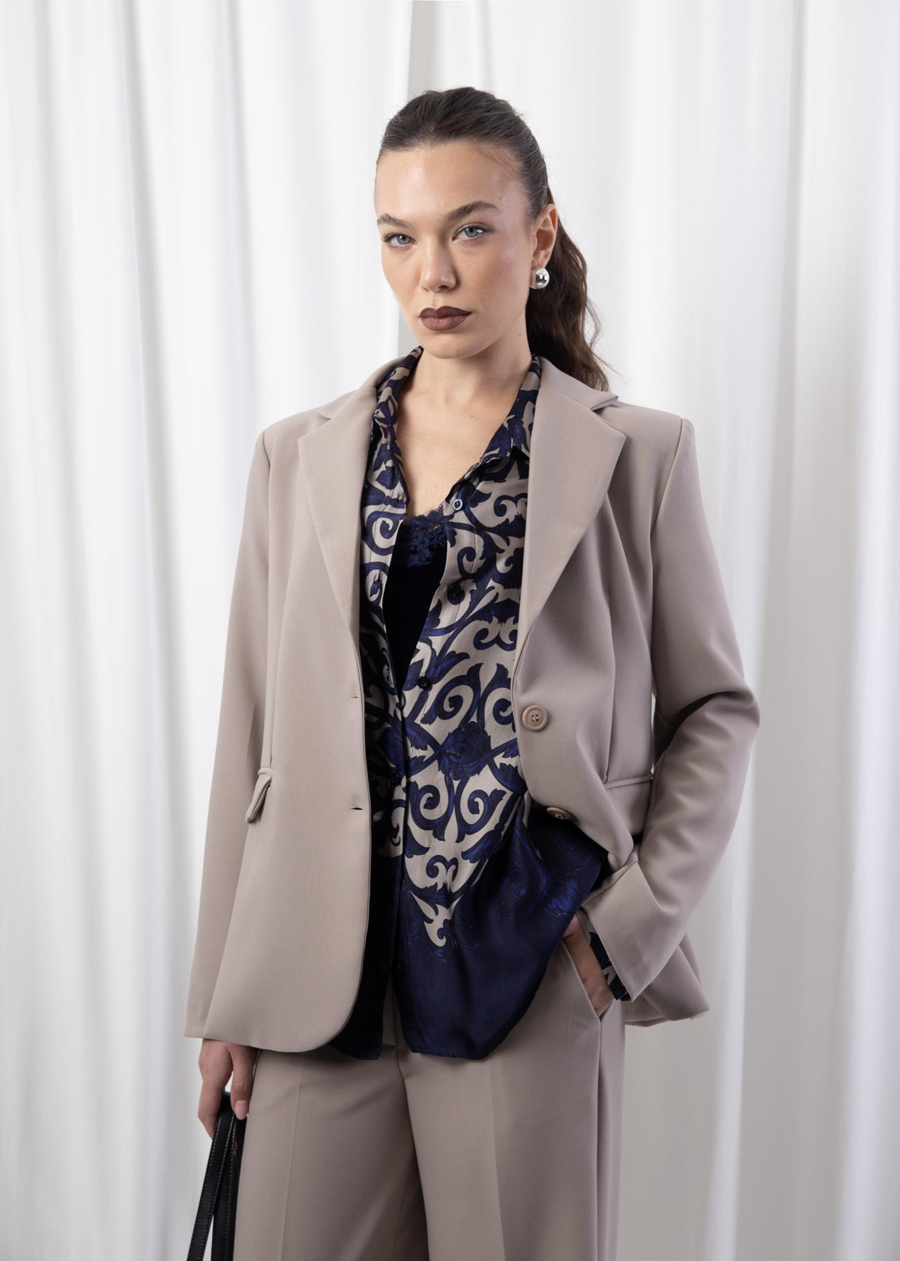 Model wearing a taupe single-breasted blazer with flap pockets, tonal buttons, and padded shoulders, photographed against a white backdrop.