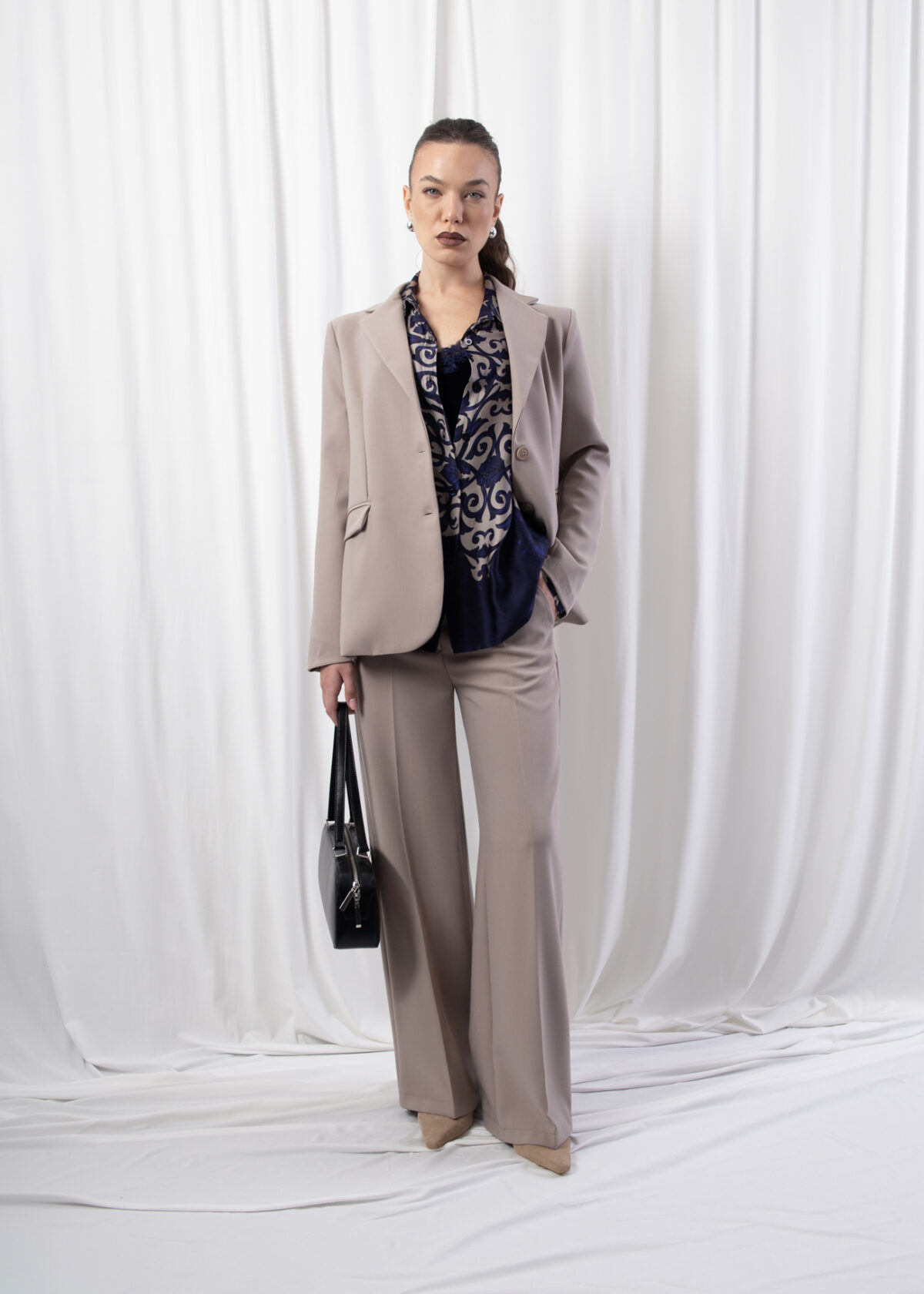 Model wearing high-rise wide-leg trousers in a neutral beige tone, featuring pressed creases and elasticated back waistband, styled with matching blazer.