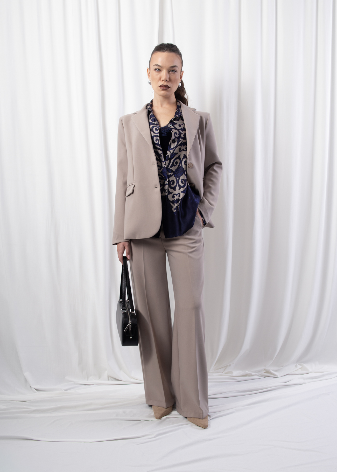Model wearing high-rise wide-leg trousers in a neutral beige tone, featuring pressed creases and elasticated back waistband, styled with matching blazer.