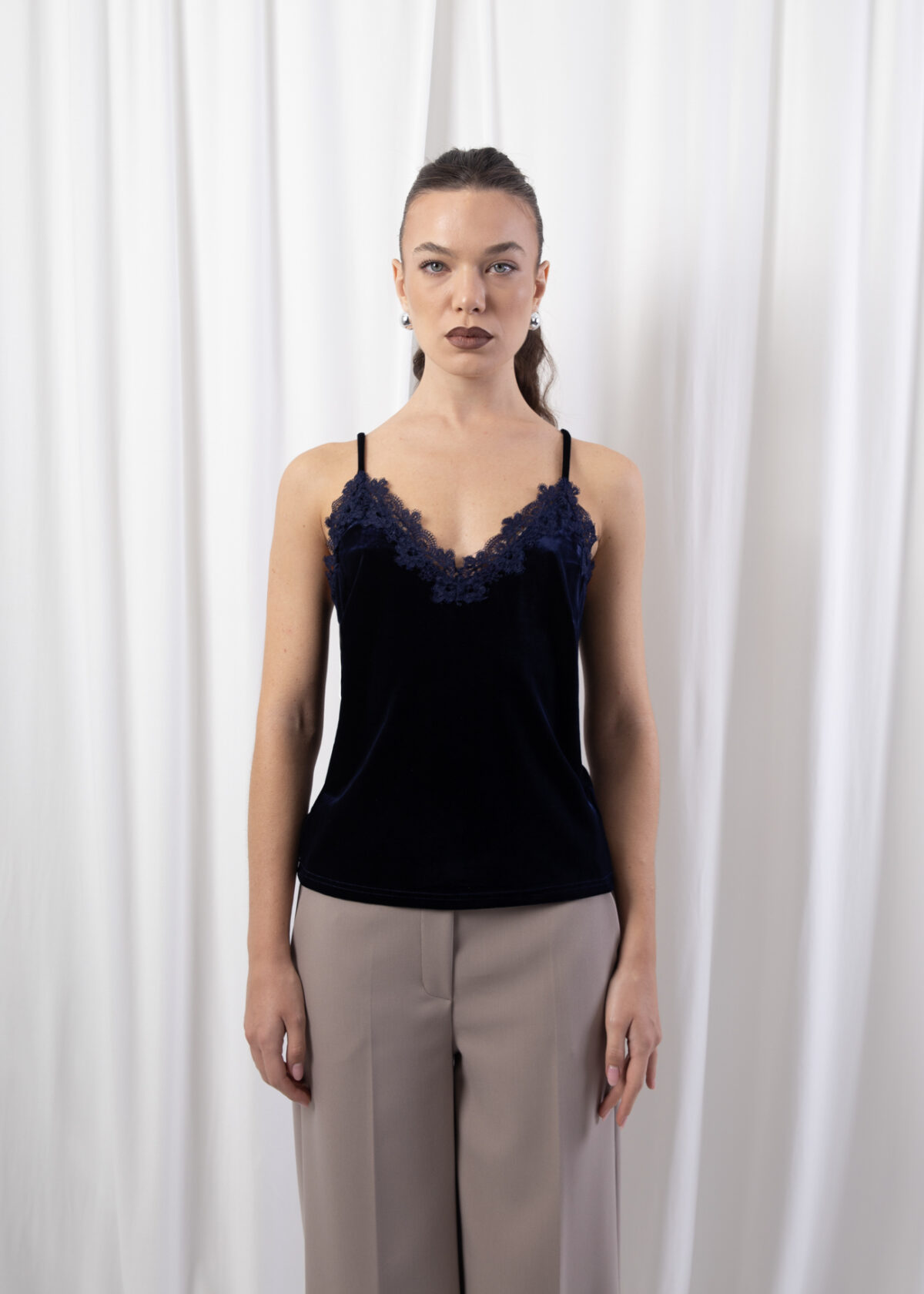 Model wearing a navy velvet camisole with lace-trim V-neckline and adjustable straps, styled with tailored trousers against a white backdrop.