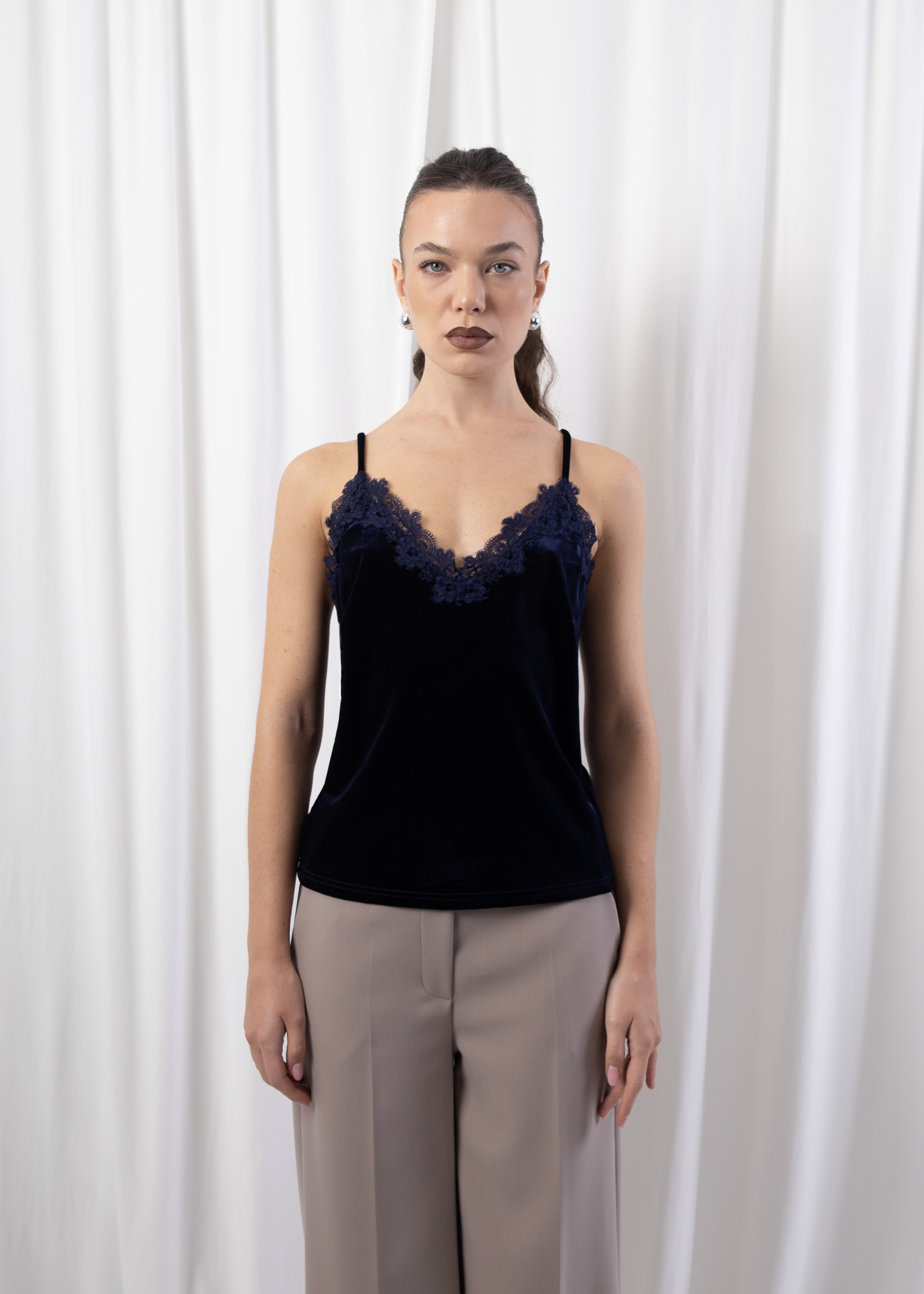 Model wearing a navy velvet camisole with lace-trim V-neckline and adjustable straps, styled with tailored trousers against a white backdrop.