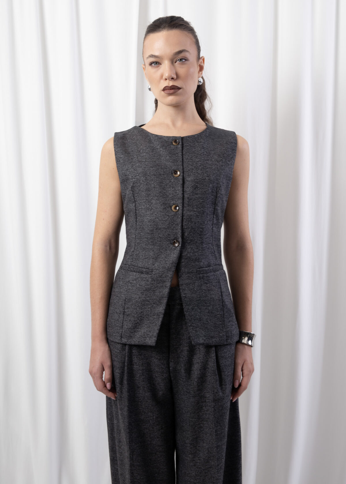 Model wearing a sleeveless tailored gilet with round neckline, front buttons, and flap pockets, styled with matching wide-leg trousers against a white backdrop.