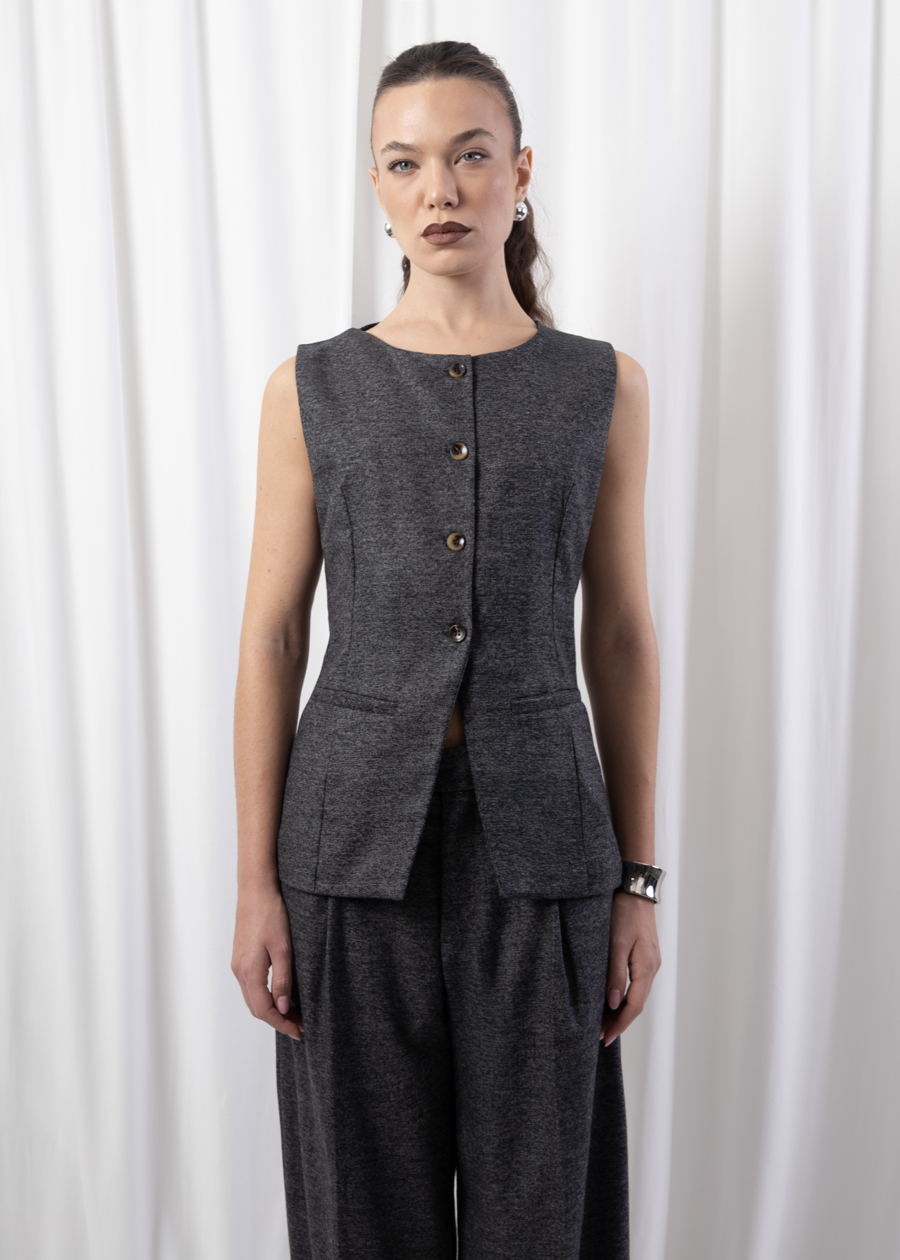 Model wearing a sleeveless tailored gilet with round neckline, front buttons, and flap pockets, styled with matching wide-leg trousers against a white backdrop.