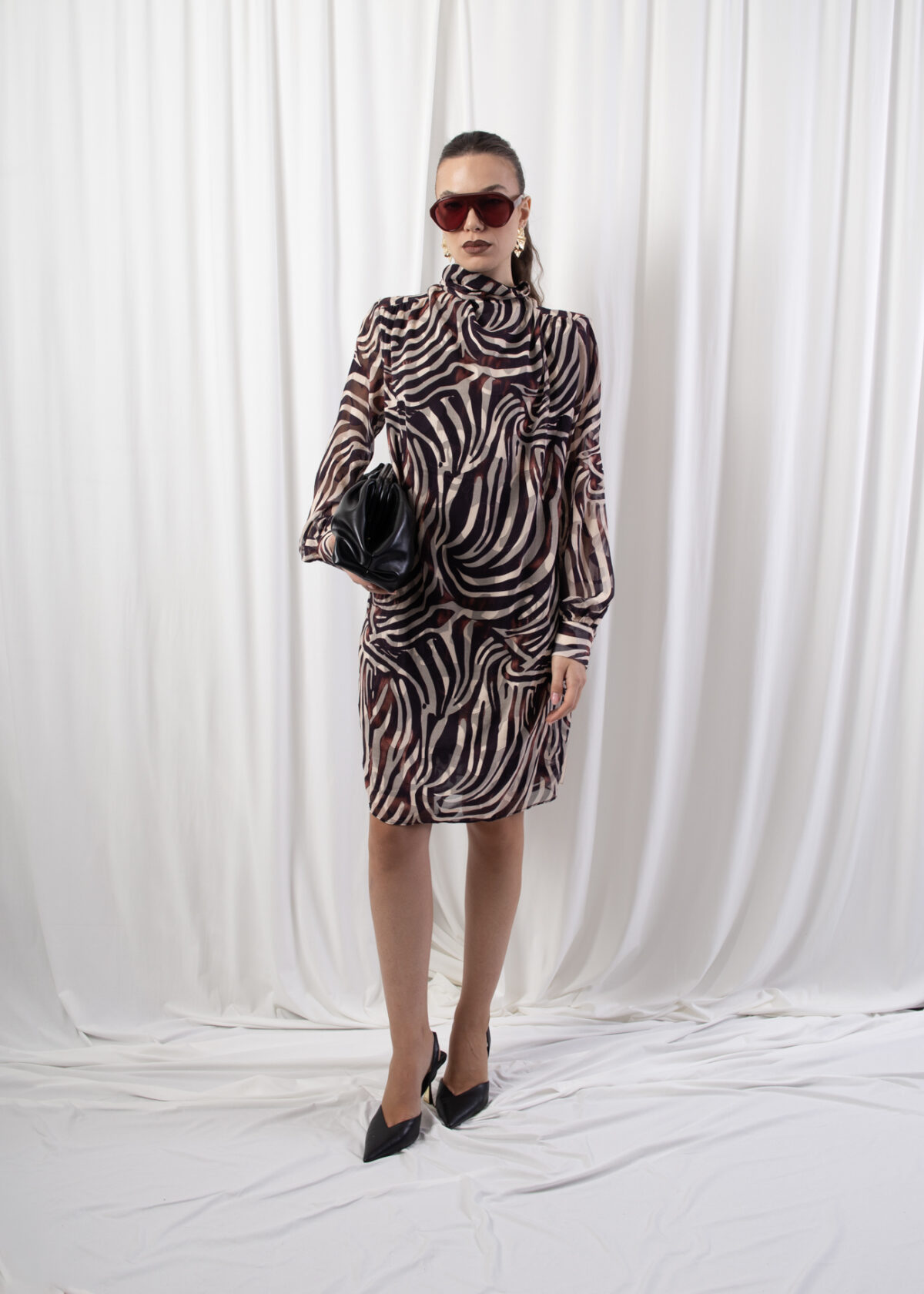 Model wearing a high neck long sleeve dress with buttoned cuffs and back button closure, styled with accessories against a white draped background.