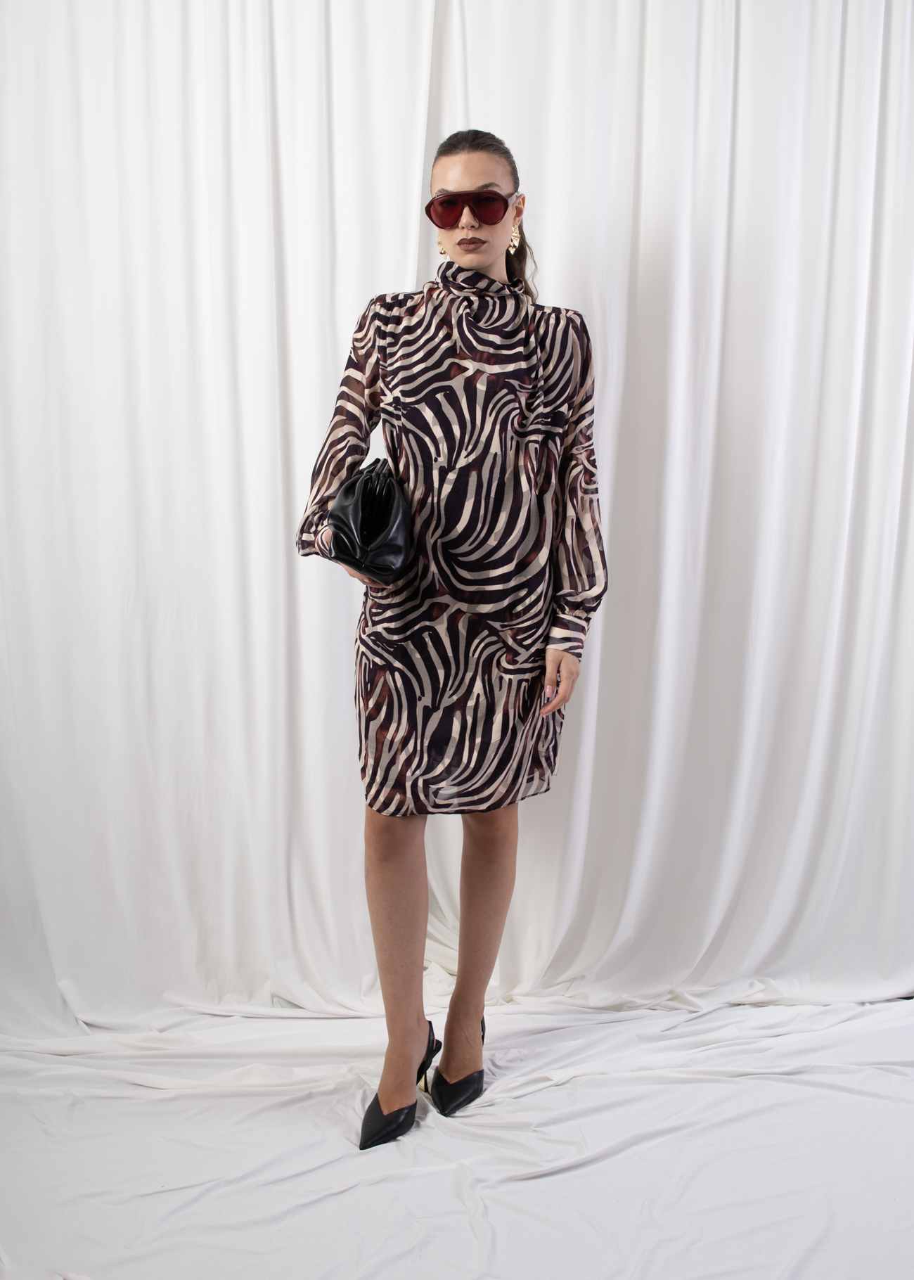 Model wearing a high neck long sleeve dress with buttoned cuffs and back button closure, styled with accessories against a white draped background.