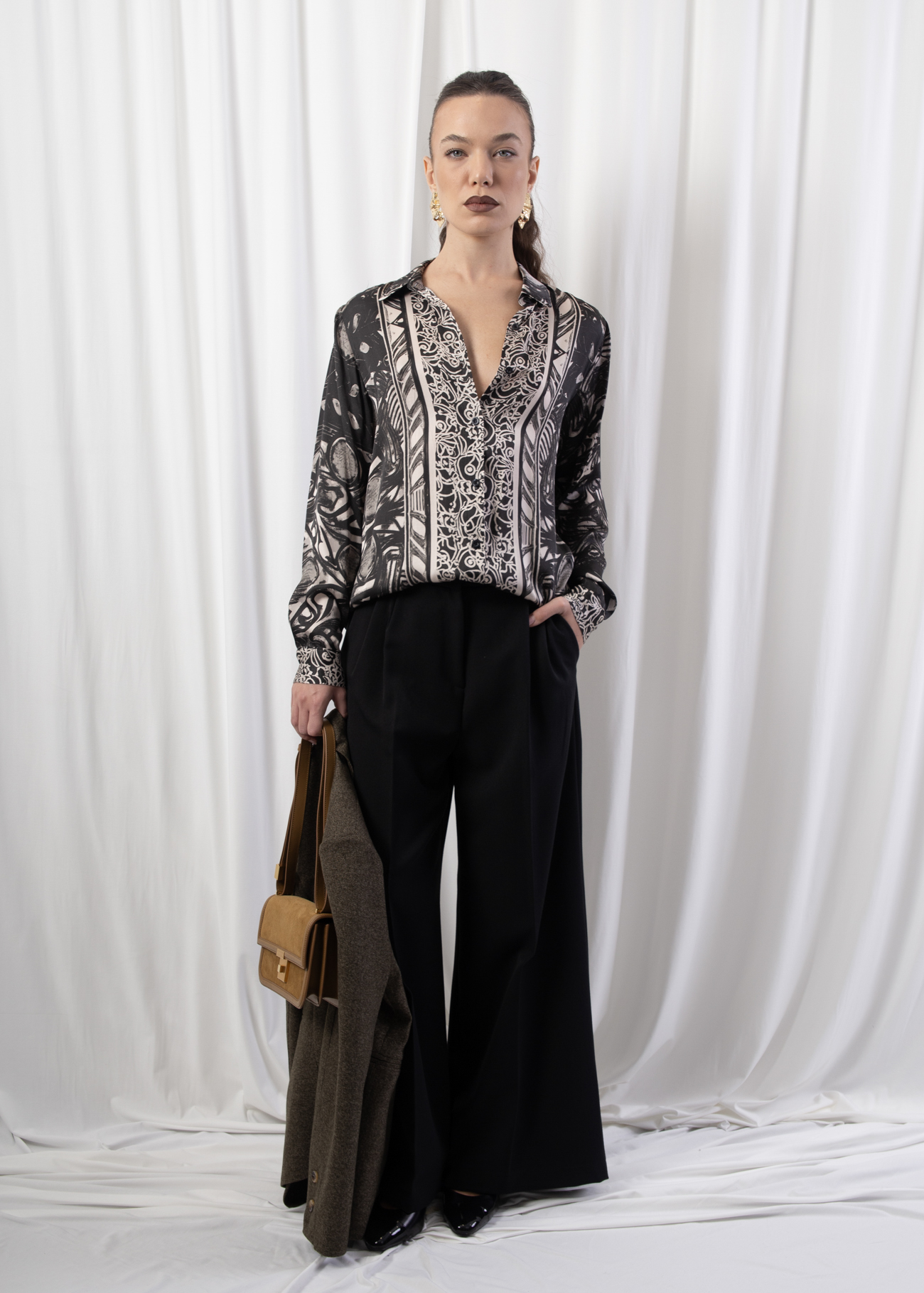 Model wearing wide-leg tailored trousers with high-rise waist and front pleats, styled with a printed blouse and heels, photographed against a white draped background.