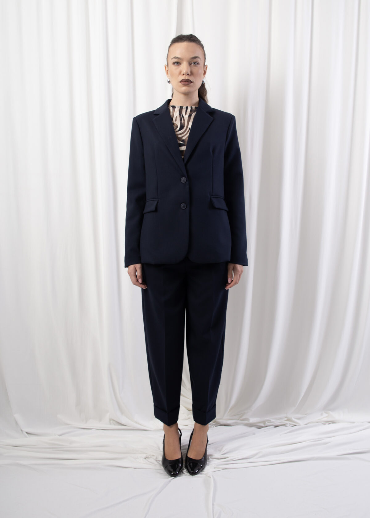 Model wearing a single-breasted tailored blazer with notched lapel, two-button closure, and flap welt pockets, photographed against a draped white backdrop.