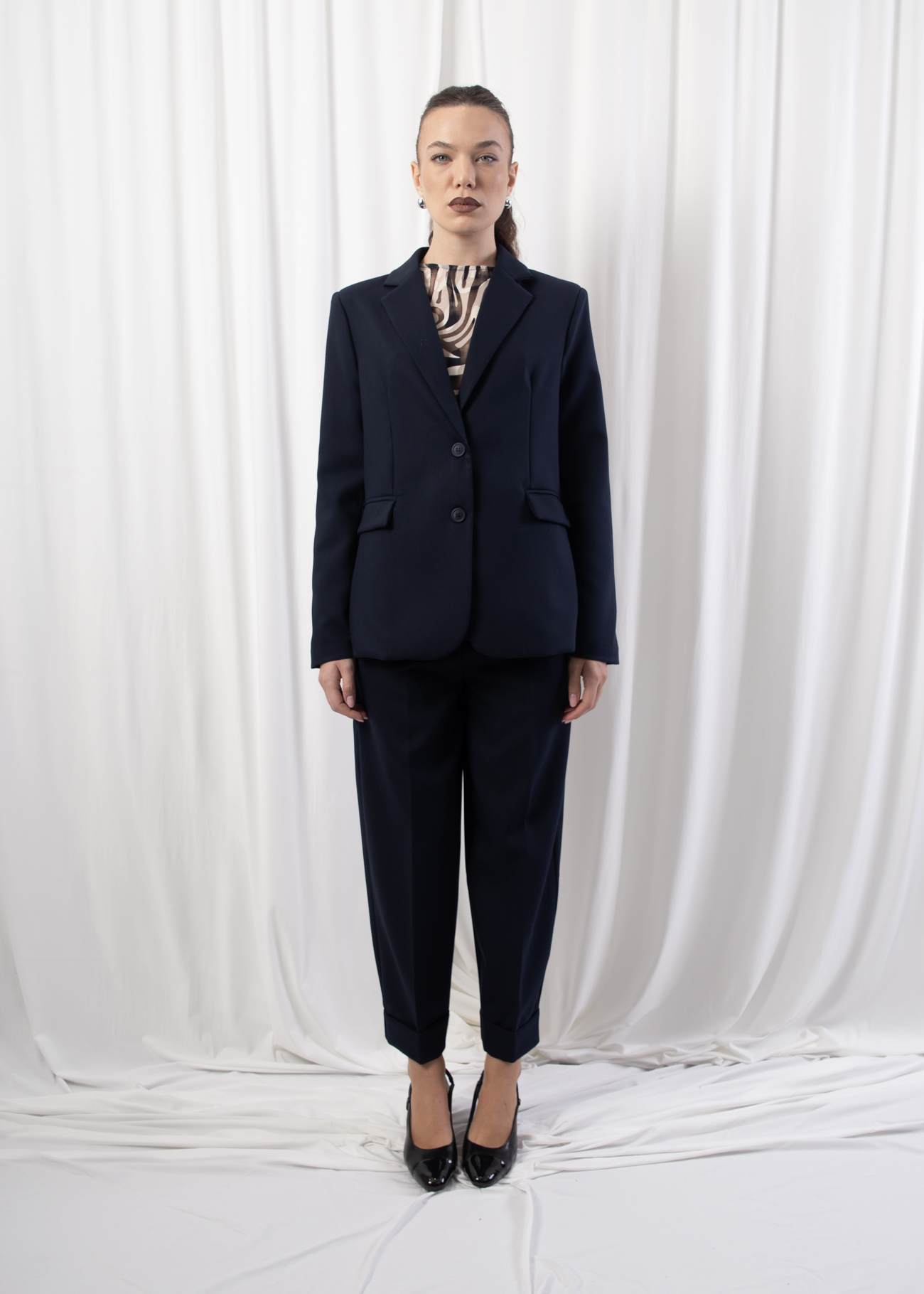 Model wearing a single-breasted tailored blazer with notched lapel, two-button closure, and flap welt pockets, photographed against a draped white backdrop.