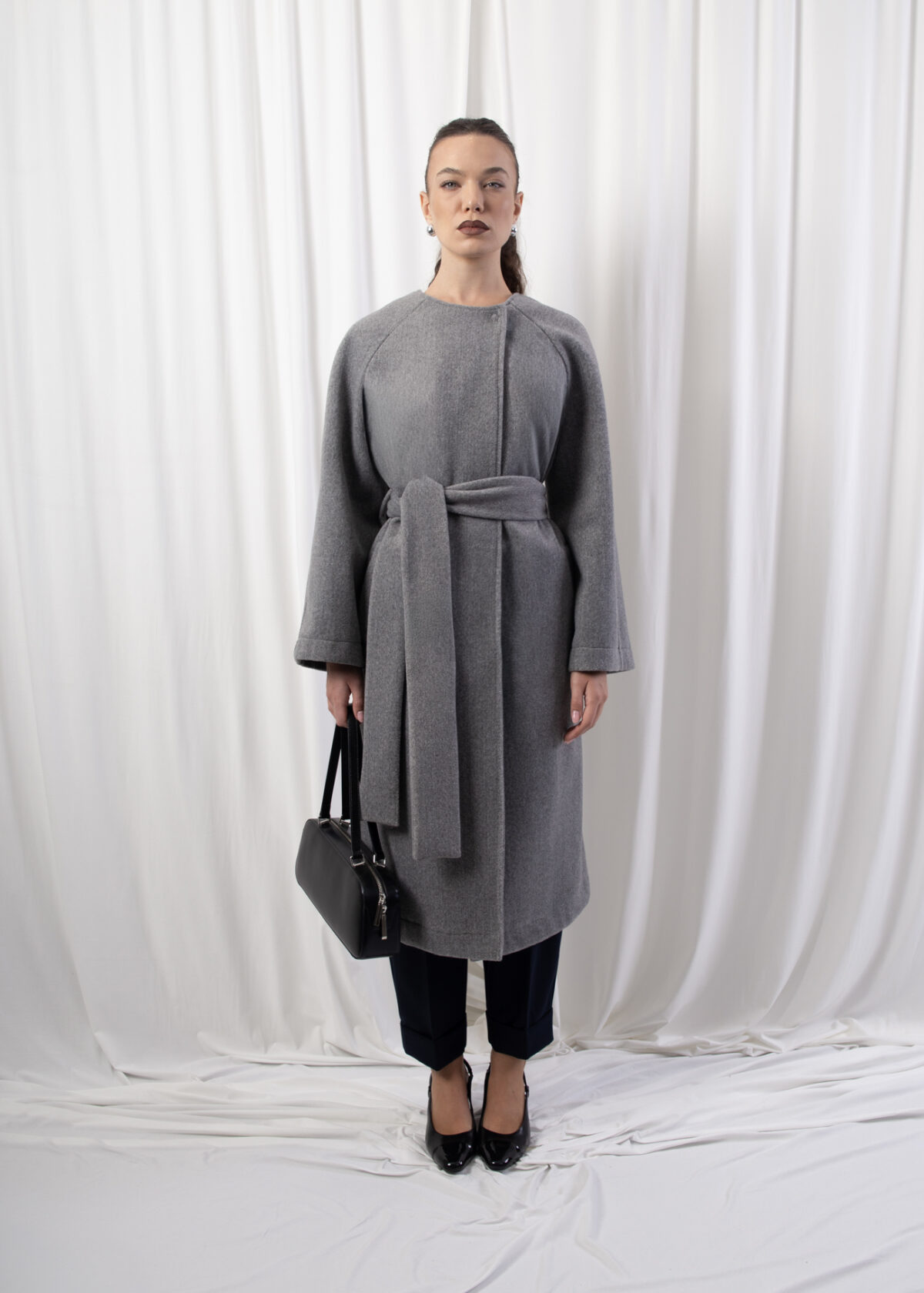 Model wearing a longline belted coat with raglan sleeves and concealed button closure, photographed against a white backdrop.