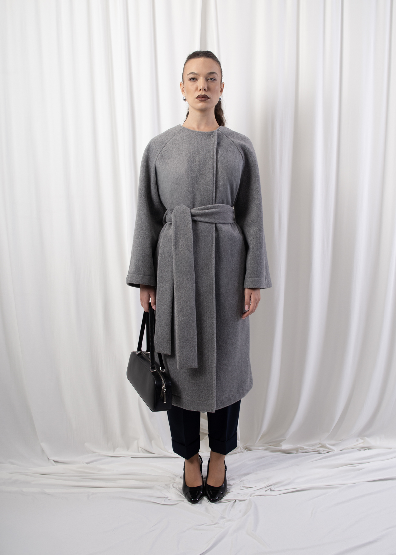 Model wearing a longline belted coat with raglan sleeves and concealed button closure, photographed against a white backdrop.