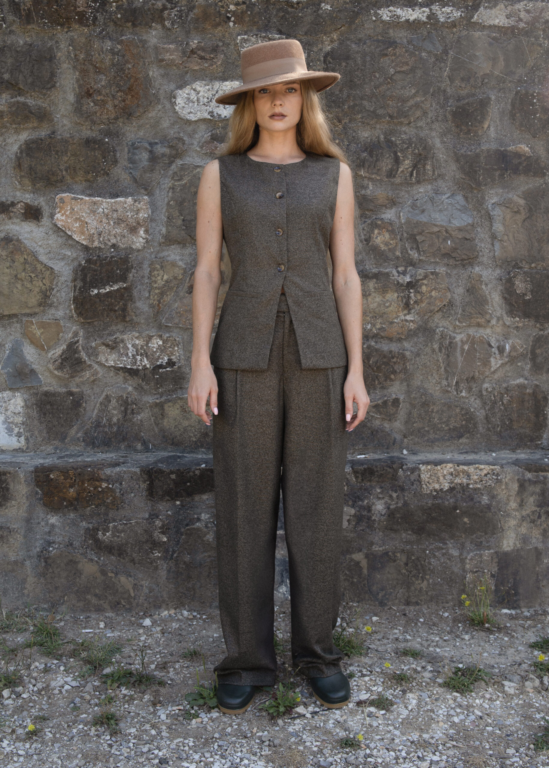 Model wearing wide-leg tailored trousers with front pleats and side pockets, styled with a sleeveless vest and hat, photographed outdoors against a stone wall.