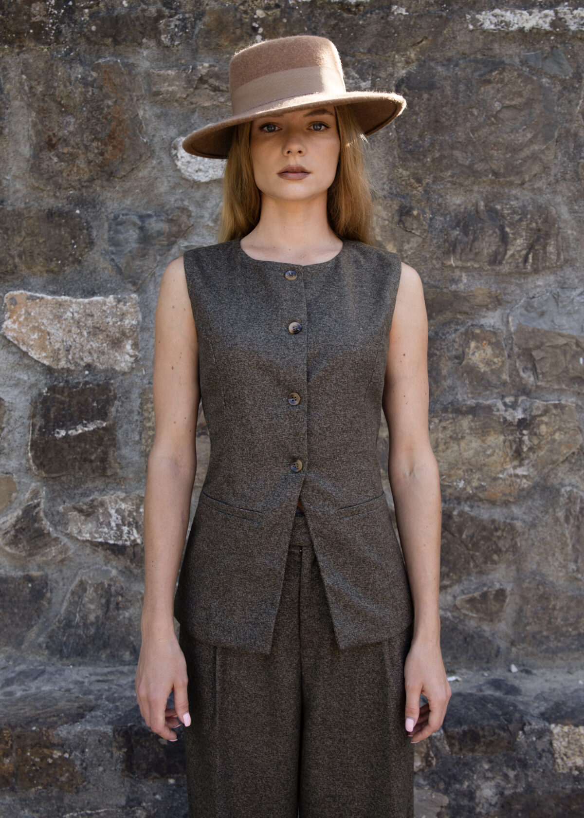 Model wearing a structured sleeveless vest with button closure and elongated silhouette, styled with matching trousers and a hat, photographed outdoors against a stone wall.
