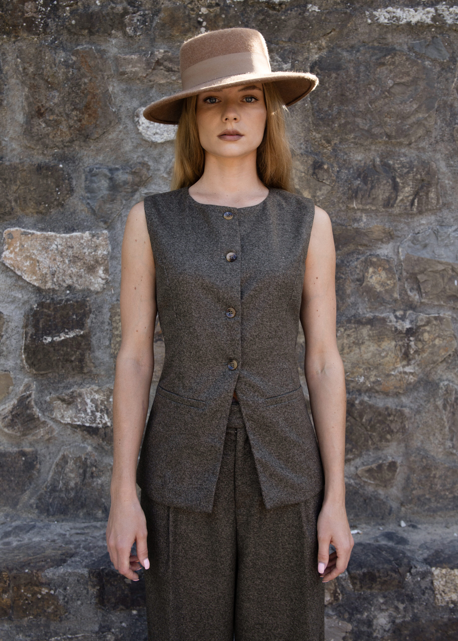 Model wearing a structured sleeveless vest with button closure and elongated silhouette, styled with matching trousers and a hat, photographed outdoors against a stone wall.