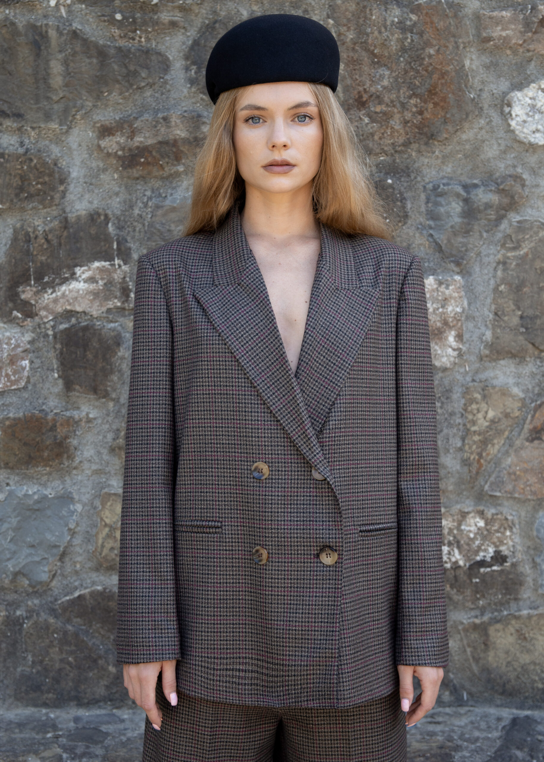 Model wearing a checked double-breasted blazer with padded shoulders, peak lapels, and flap pockets, styled with matching tailored shorts, photographed outdoors against a stone wall.