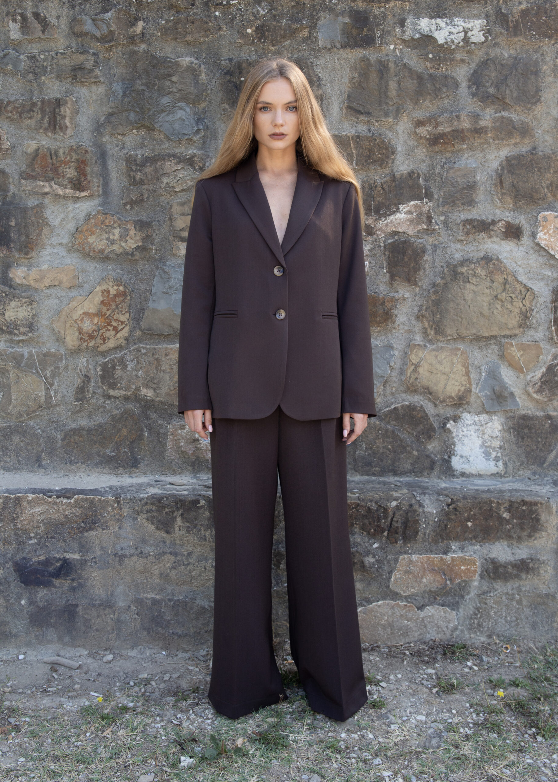 Model wearing high-rise wide-leg trousers with front pleats, back flap pockets, and pressed crease, styled with a matching blazer in outdoor setting.