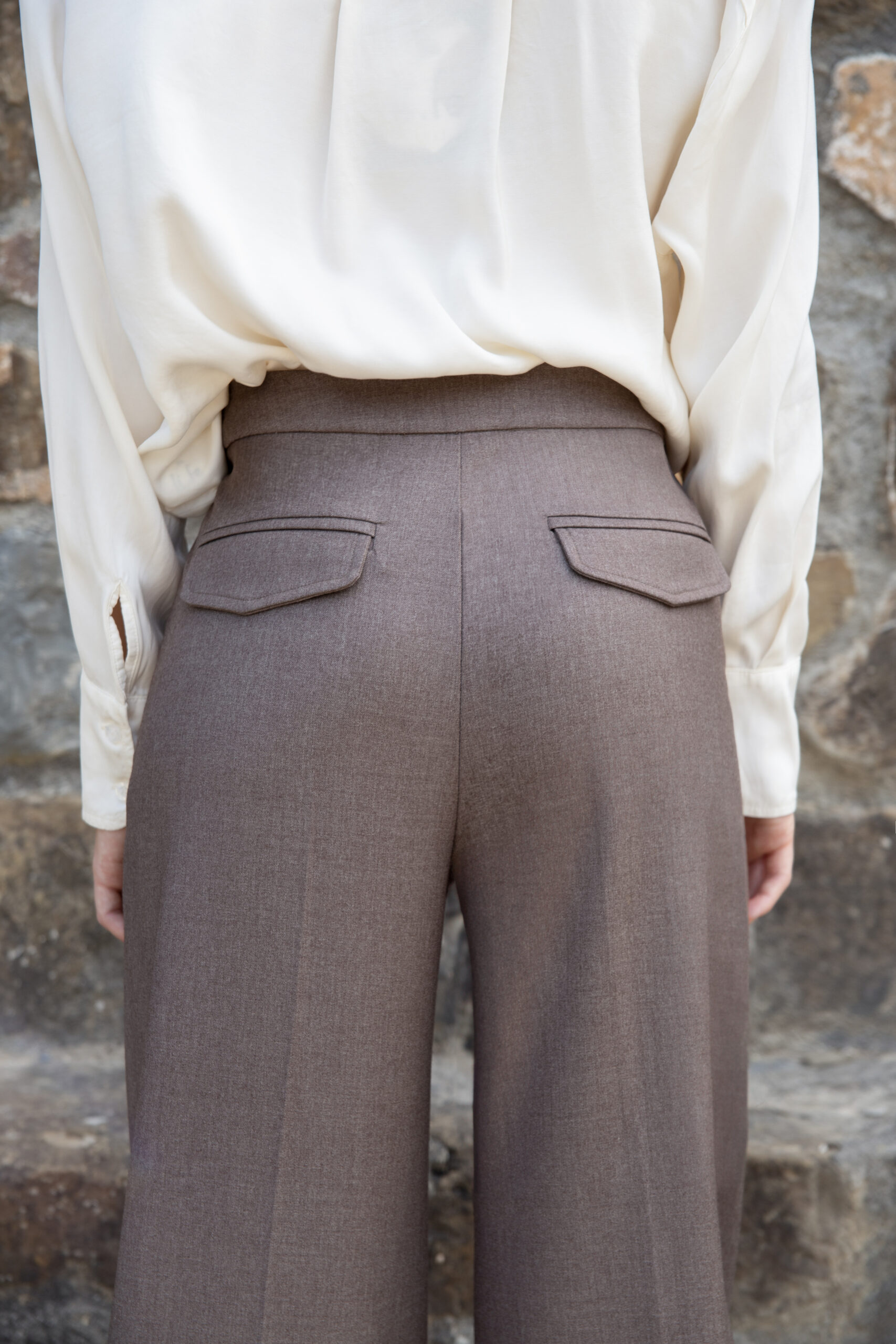 Model wearing high-rise wide-leg tailored trousers with front crease and back flap pockets, styled with a blazer, photographed outdoors.