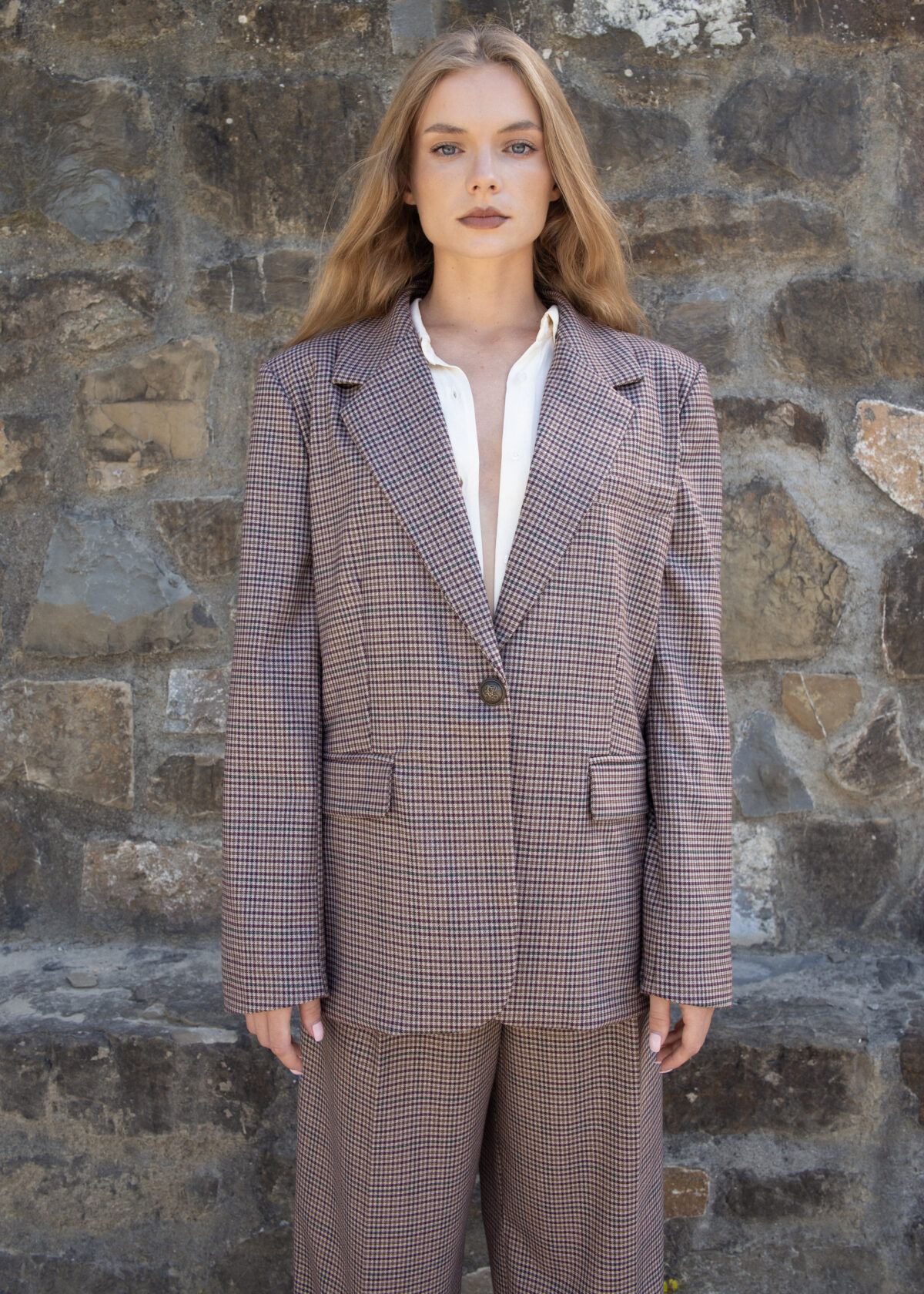 Model wearing an oversized checked blazer in brown tones with notched lapel and single button closure, styled outdoors against a stone wall.