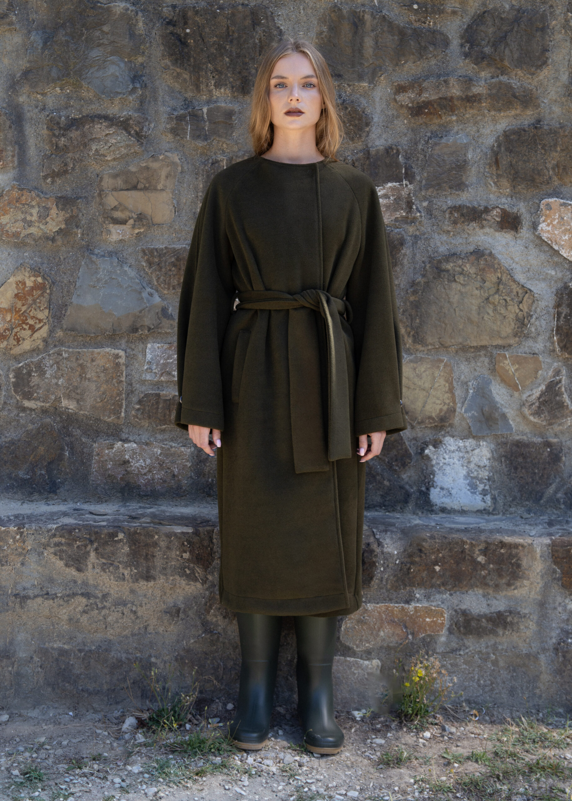 Model wearing a long wool-blend wrap coat with wide sleeves and belted waist, photographed outdoors against a stone wall and near a lake.
