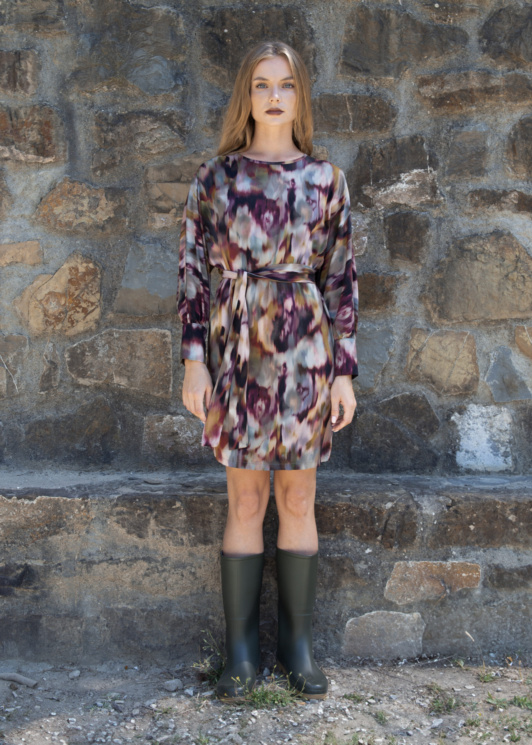 Model wearing a long-sleeve viscose dress with round neckline, removable belt, and buttoned cuffs, photographed against a stone wall.