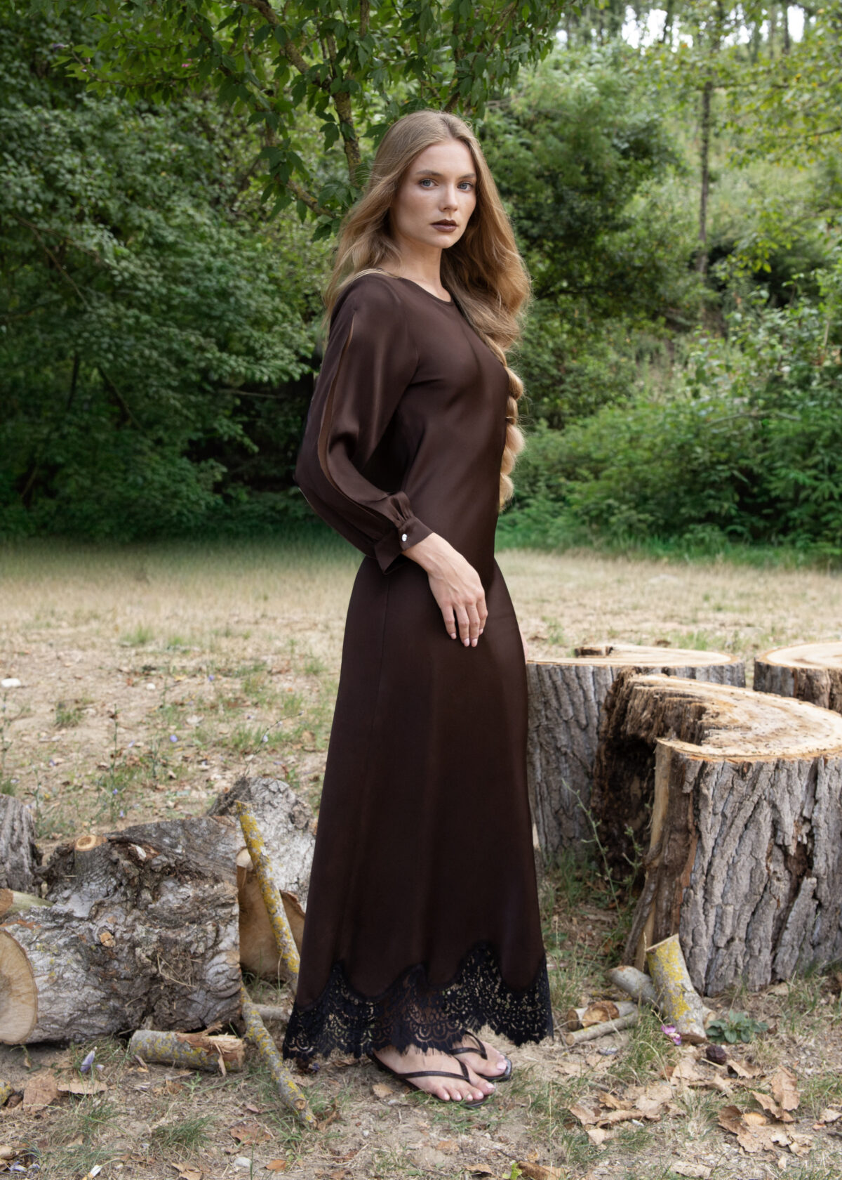 Model wearing a long brown viscose dress with sheer slit sleeves and lace hem, styled with crystal button details, photographed outdoors in natural light.