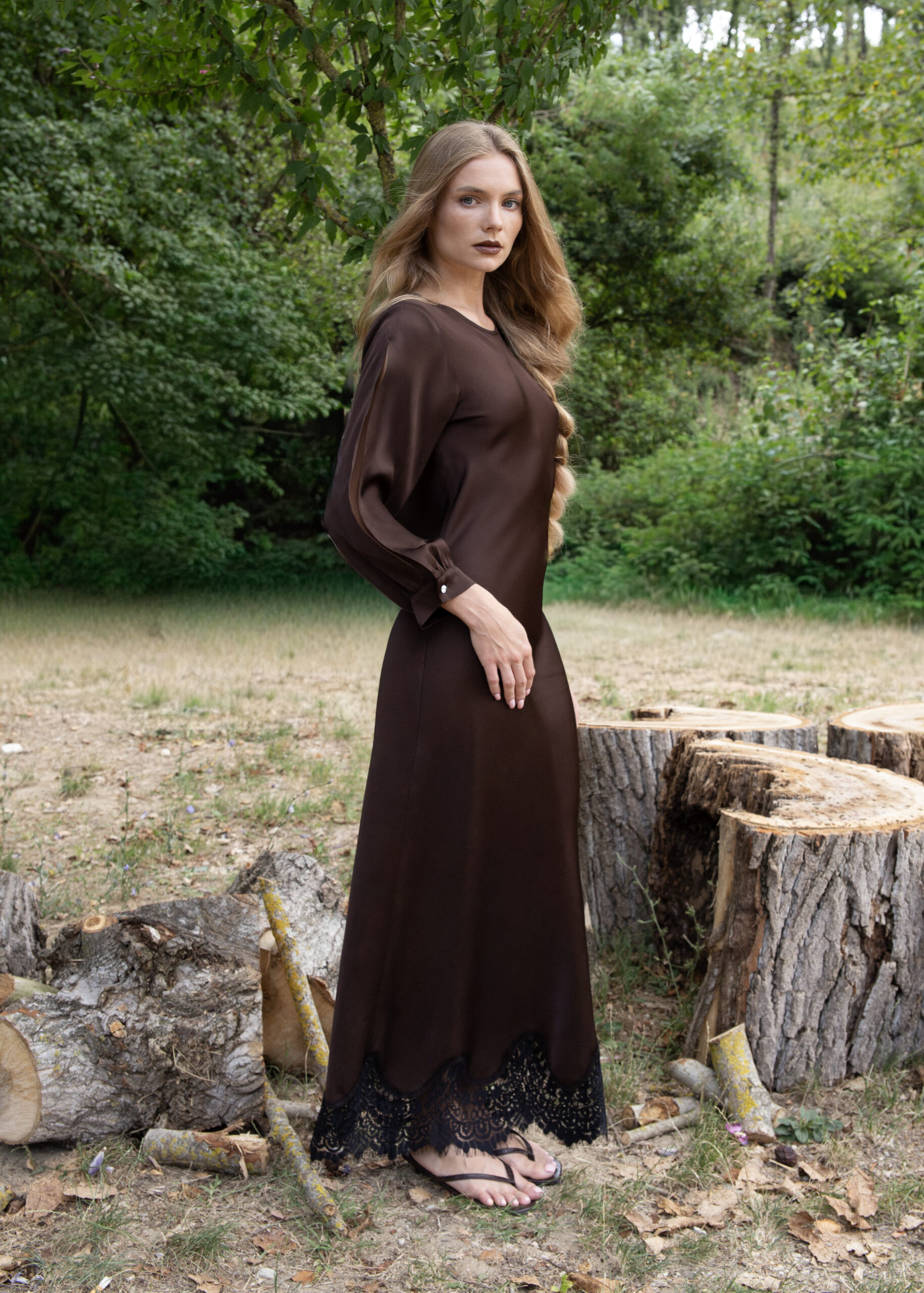 Model wearing a long brown viscose dress with sheer slit sleeves and lace hem, styled with crystal button details, photographed outdoors in natural light.